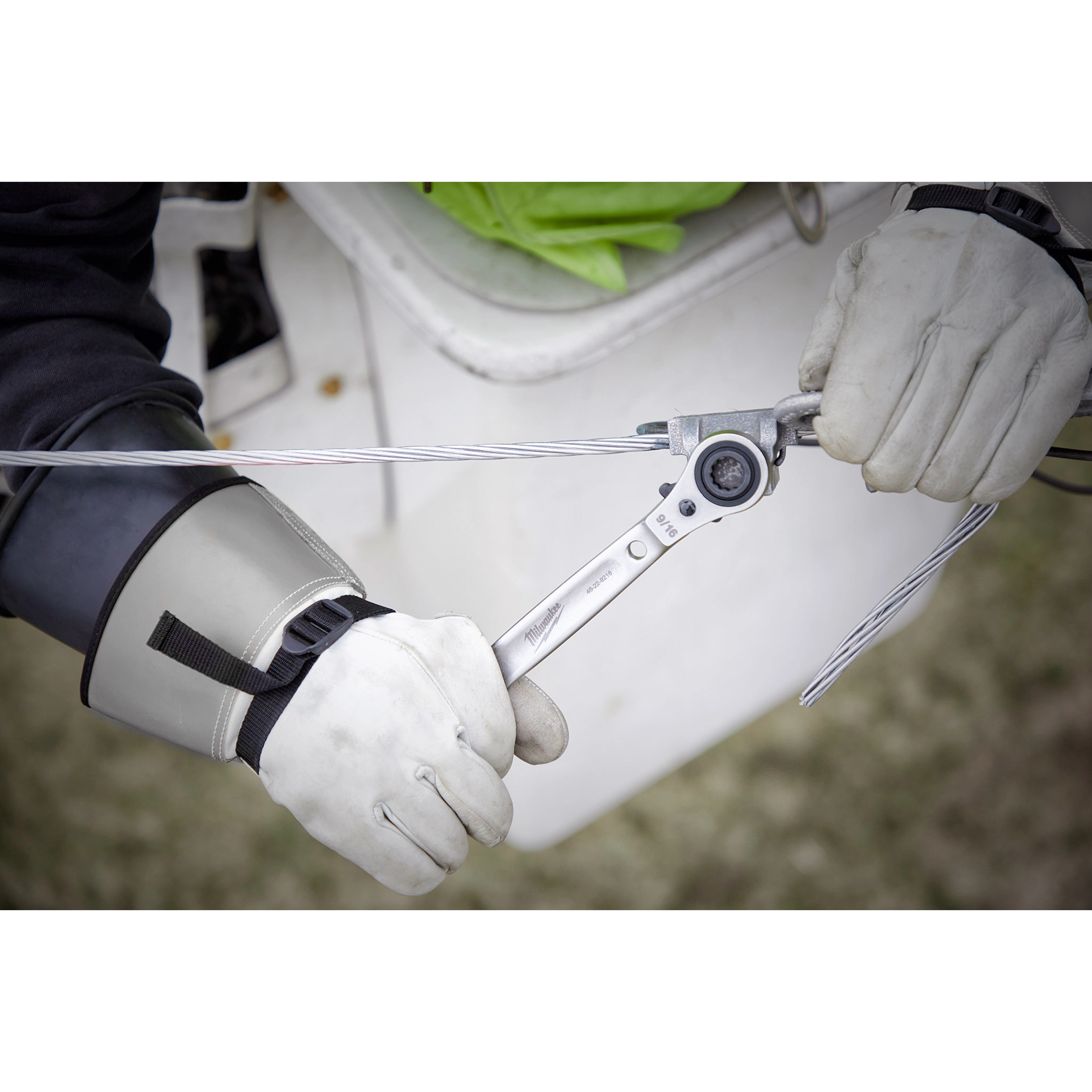 Gloved hands using Lineman's 5in1 Ratcheting Wrench on electrical cable, with a white bucket truck and safety gear visible.