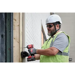 A construction worker wearing Full Frame Safety Glasses with Removable Side Shields – Silver Mirrored Anti-Scratch Lenses is using a red and black power drill to secure a screw into a wooden beam. The worker also wears a white hard hat, black gloves, and a green safety vest.