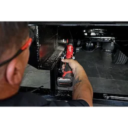 A person uses the M12 FUEL™ 1/2" Ratchet Kit to tighten a bolt in a difficult-to-reach area under a vehicle. The red and black ratchet features a battery pack and ergonomic handle.