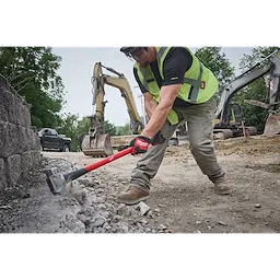 A construction worker is using an 8-lb Sledge Hammer (36” Handle) to break up concrete on a job site. The hammer has a red and black handle, and excavators and trucks are in the background on the dirt. The worker wears gloves, a safety vest, and work boots.
