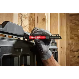 A gloved hand holding the INKZALL™ 2IN1 Deep Reach Ultra Fine Point Black Jobsite Marker is marking a dark plastic tool organizer mounted on a wooden wall. The marker has a red barrel with black details and branding displayed, highlighting its design suited for construction settings.