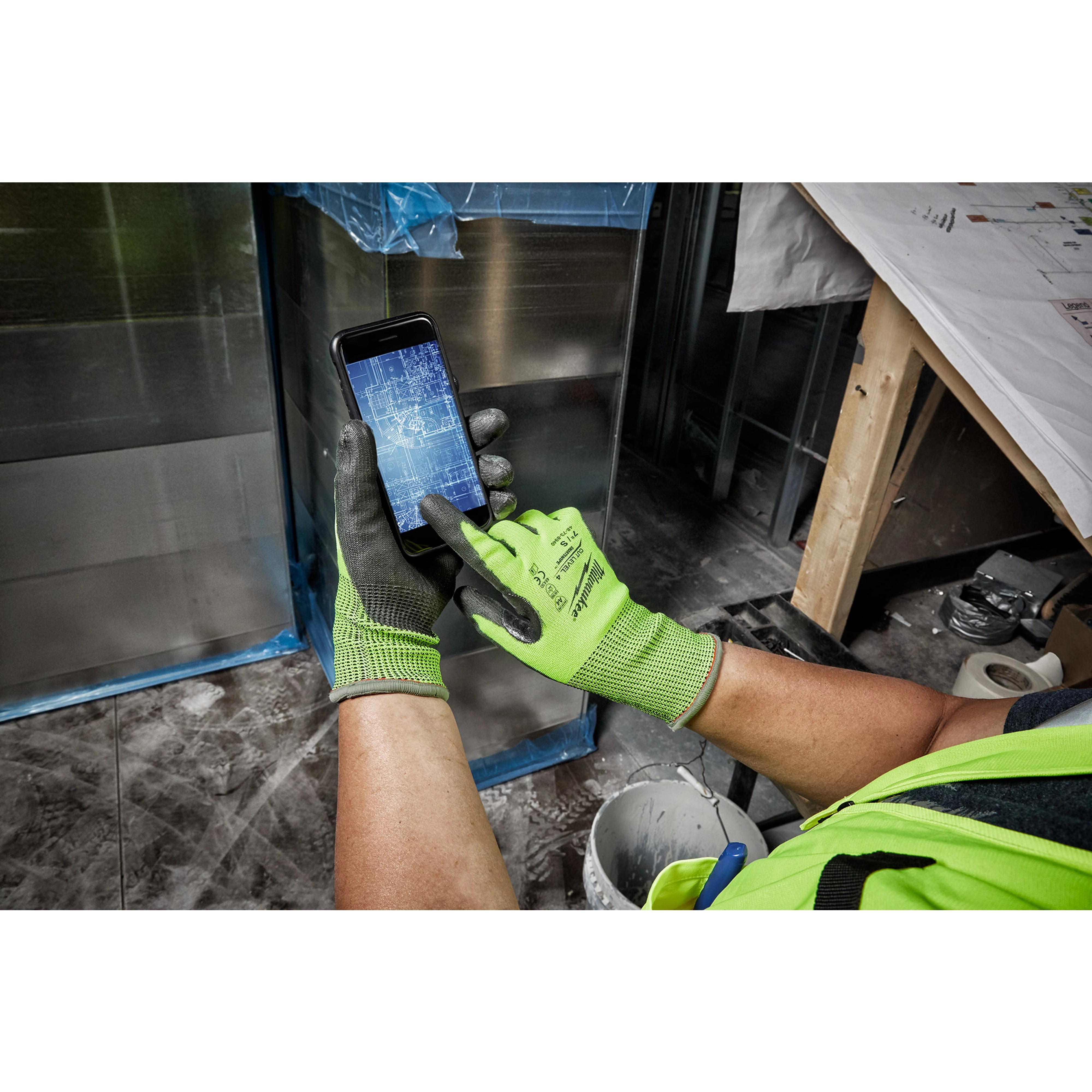 Person wearing Hi Vis Cut Gloves using a smartphone with a blueprint on the screen, in a construction worksite environment.