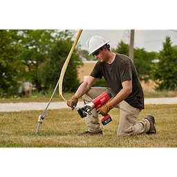 A worker using the FORCELOGIC™ M18™ 1590 ACSR Cutter Kit to cut a wire secured to a pole. The worker is wearing a white hard hat, brown short-sleeve shirt, and beige pants, while kneeling on grass.