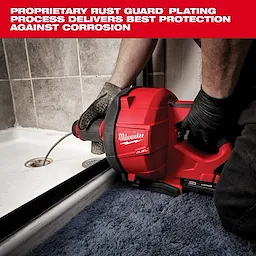 A Milwaukee Fuel drain cleaner is in use, with a worker's gloved hands guiding the cable into a shower drain. The red tool is prominently labeled with the brand name and is designed to prevent corrosion with its rust guard plating. The area surrounding the drain is tiled and has a blue carpet.