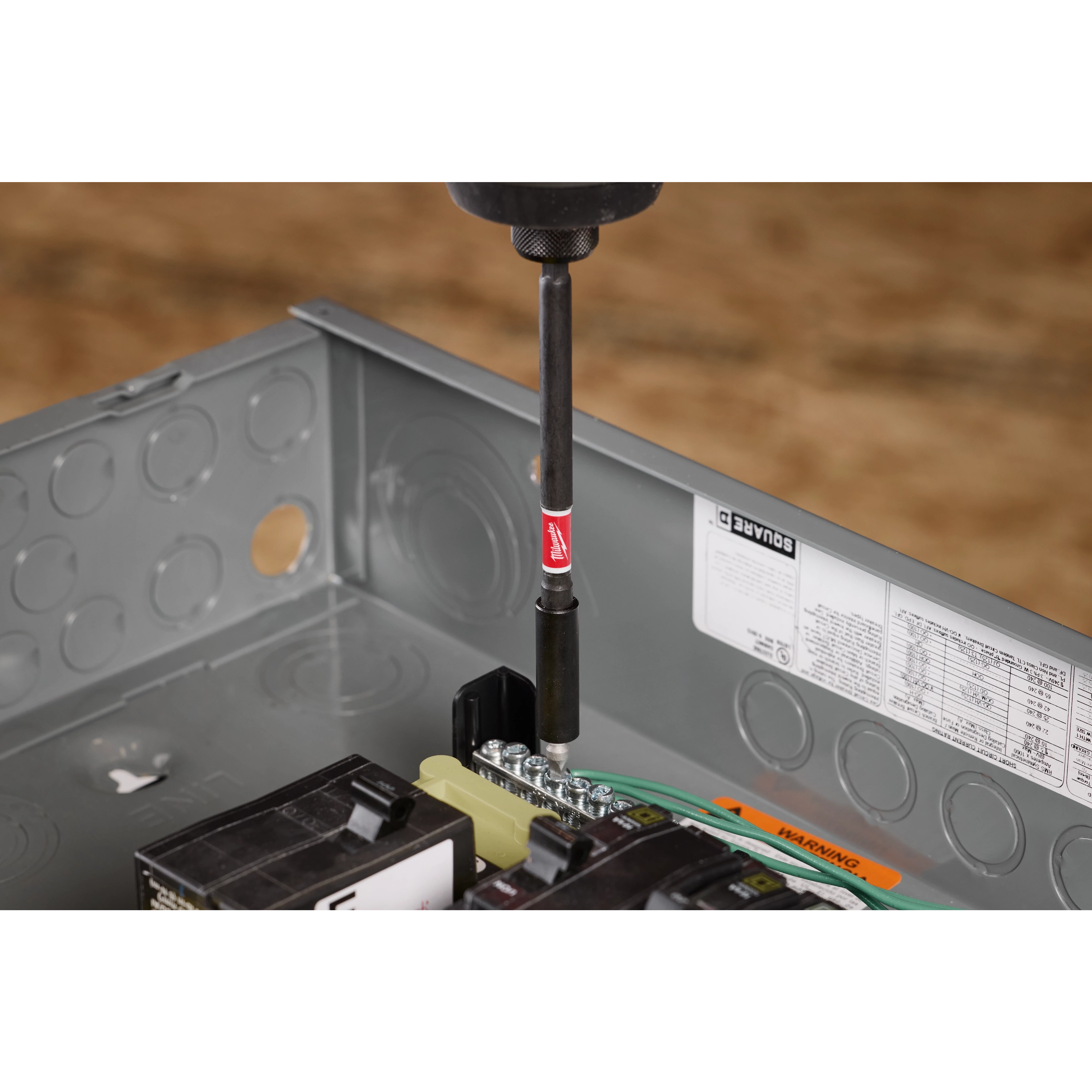 A SHOCKWAVE™ 6" Impact Magnetic Bit Holder is shown in use, attached to a drill and securing a screw into an electrical box. The magnetic bit holder is black with a red band and the SHOCKWAVE™ logo. Various tools and components are visible within the electrical box.