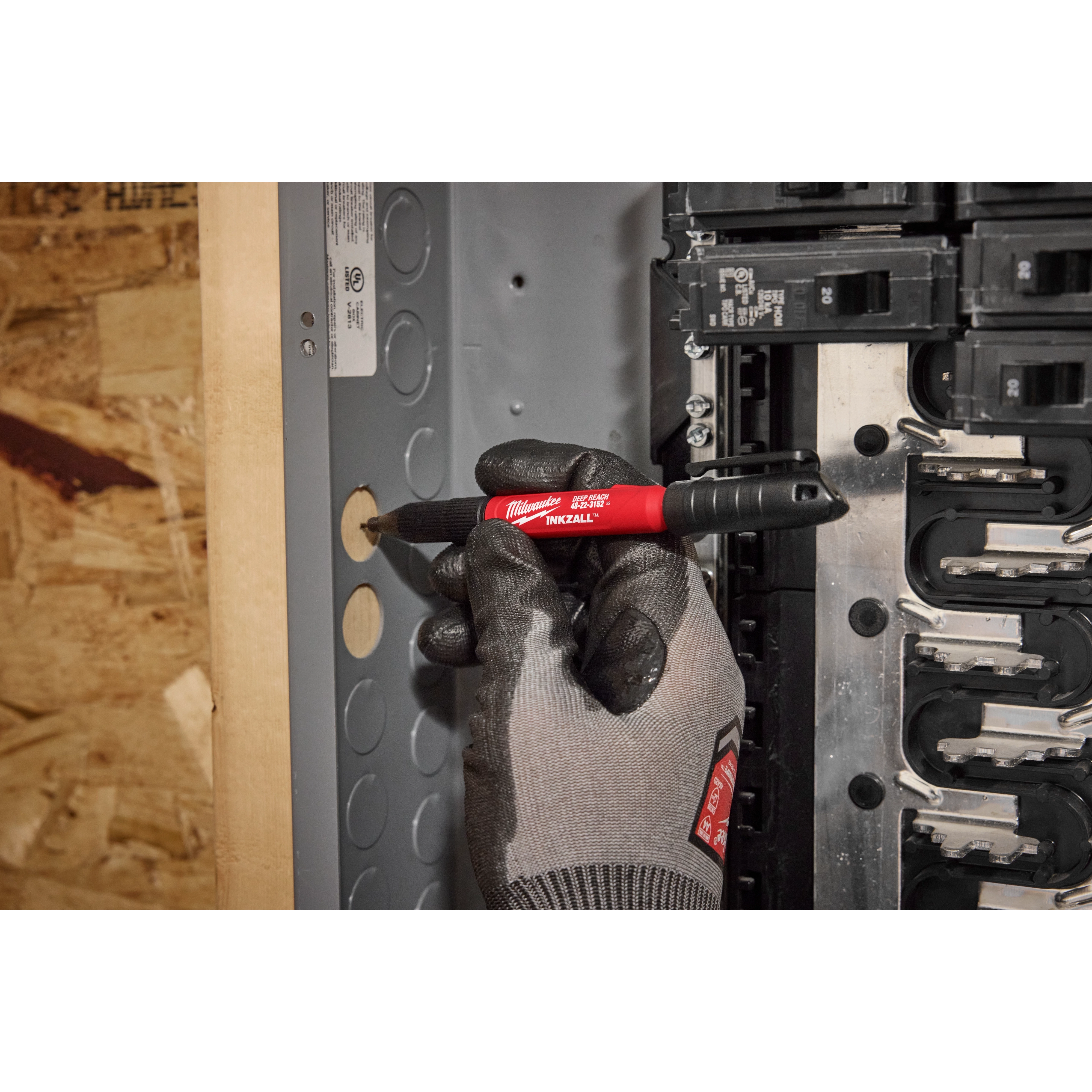 A gloved hand holds the INKZALL™ 2IN1 Deep Reach Ultra Fine Point Black Jobsite Marker, marking a hole on an electrical panel with precision. The marker’s red body and black cap are visible, while the surrounding workspace shows wood and metal materials.