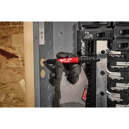 A gloved hand holds the INKZALL™ 2IN1 Deep Reach Ultra Fine Point Black Jobsite Marker, marking a hole on an electrical panel with precision. The marker’s red body and black cap are visible, while the surrounding workspace shows wood and metal materials.