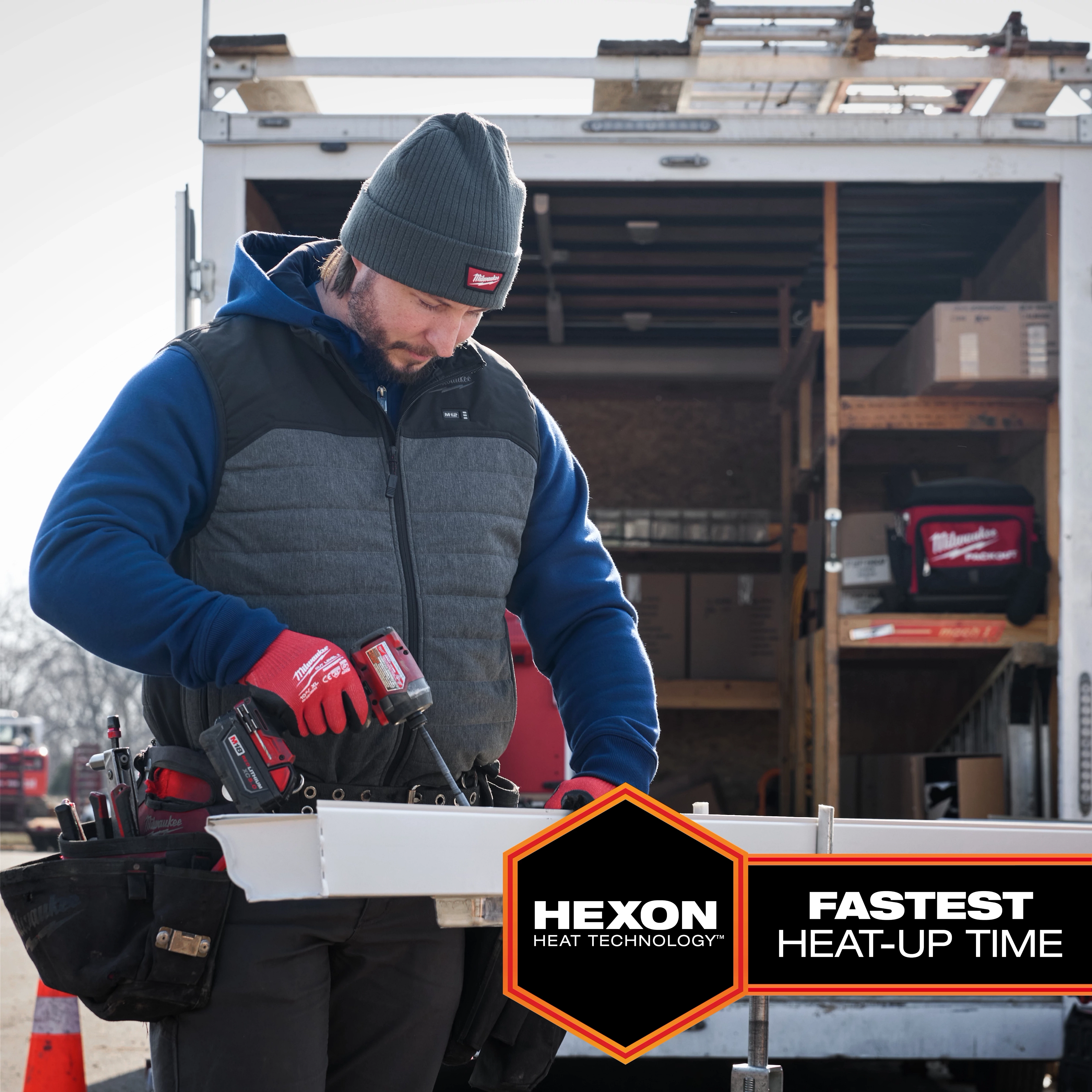 A construction worker wears an M12 Heated AXIS Vest while working with a power tool outside a trailer.