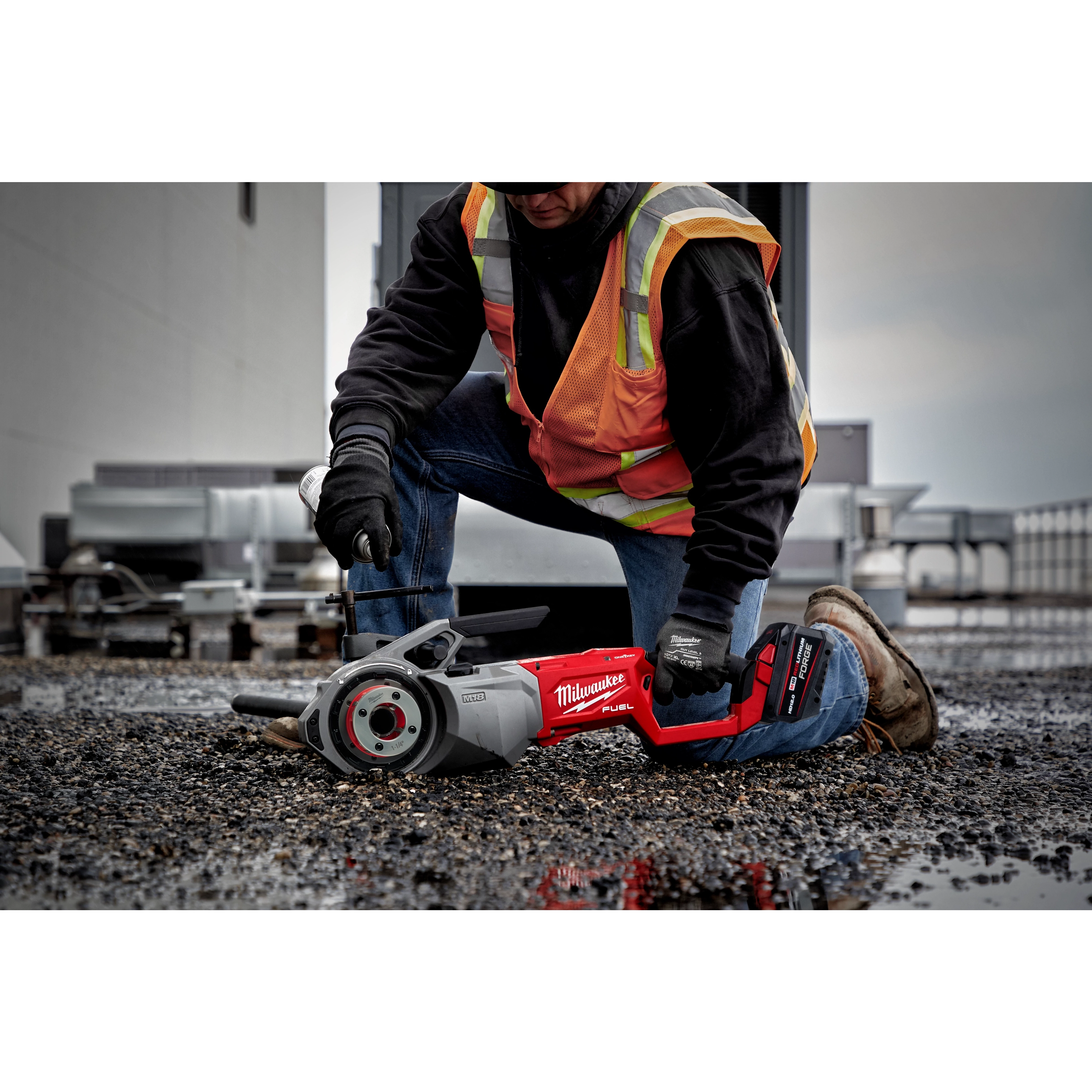 A person in a safety vest and gloves is using a Milwaukee M18 FUEL™ Pipe Threader with ONEKEY on a gravel surface, next to industrial equipment.