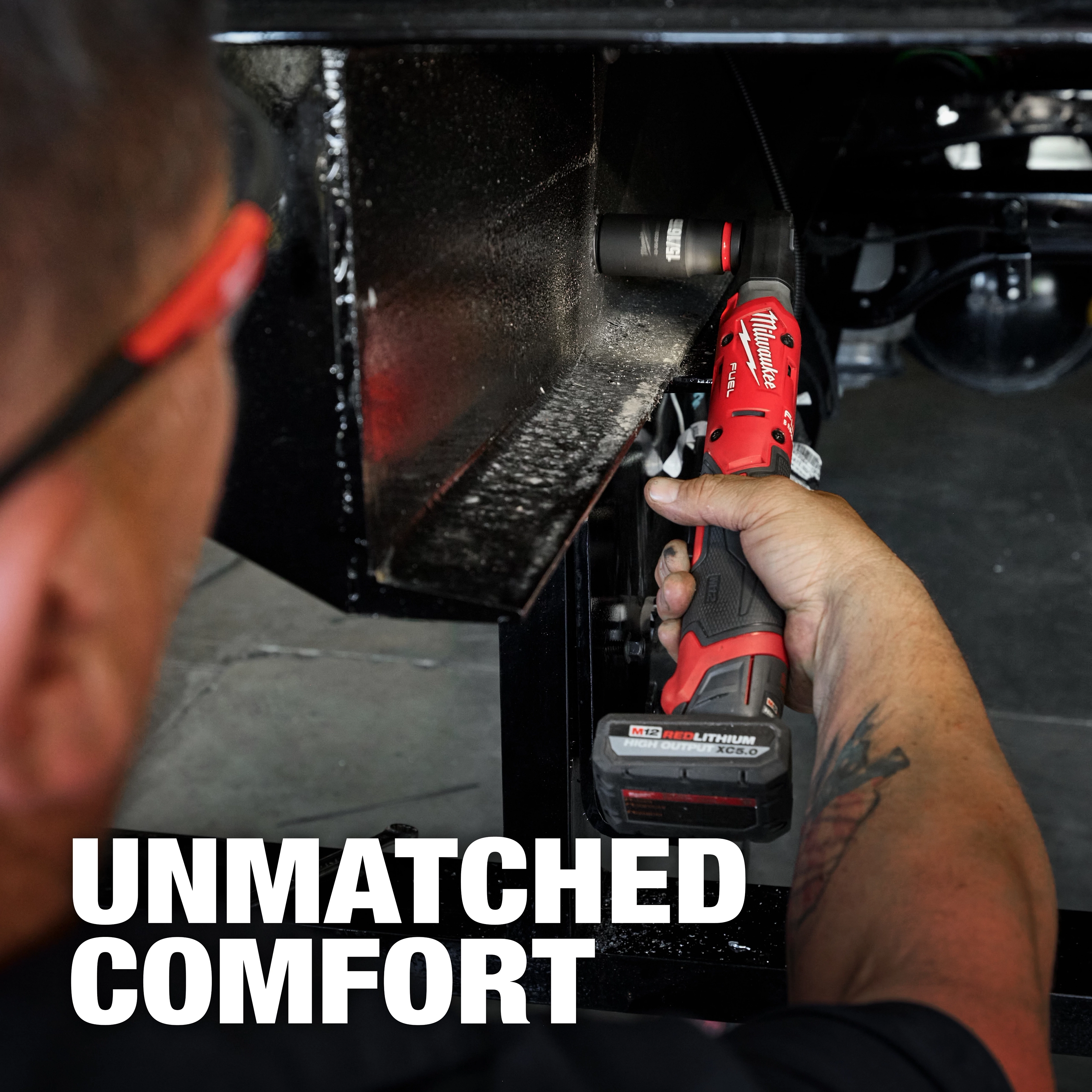A person uses the M12 FUEL™ 1/2" Ratchet to tighten a bolt in a tight space. The tool is red and black, featuring the text "UNMATCHED COMFORT" in large white letters.