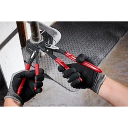 Two gloved hands using 10" Pliers Wrench with red and black handles to grip and twist a pipe. The tool is in use in a workshop environment.