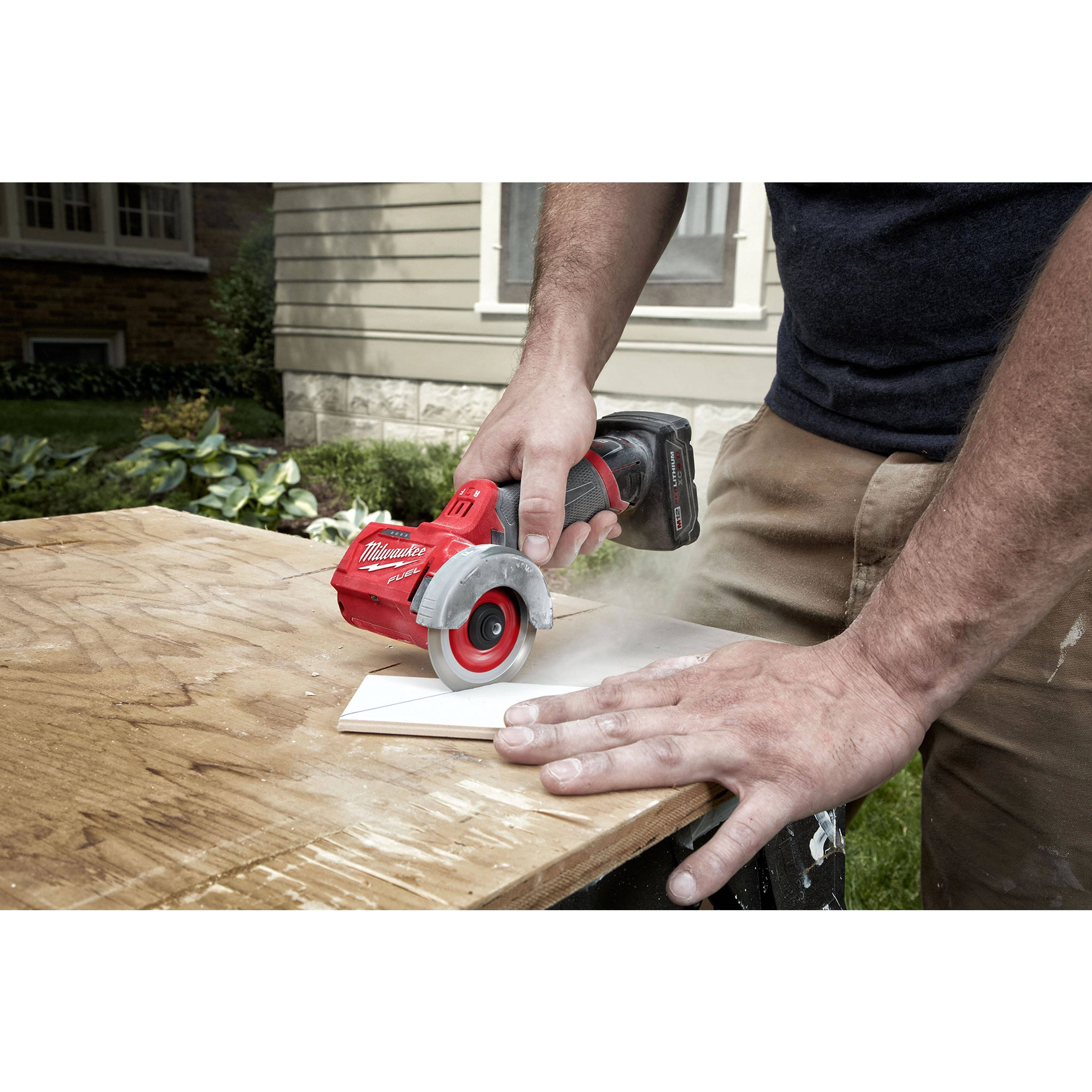 Person using the M12 FUEL 3" Compact Cut Off Tool to cut a piece of wood on a workbench outdoors. House visible in the background.