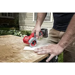 Person using the M12 FUEL 3" Compact Cut Off Tool to cut a piece of wood on a workbench outdoors. House visible in the background.