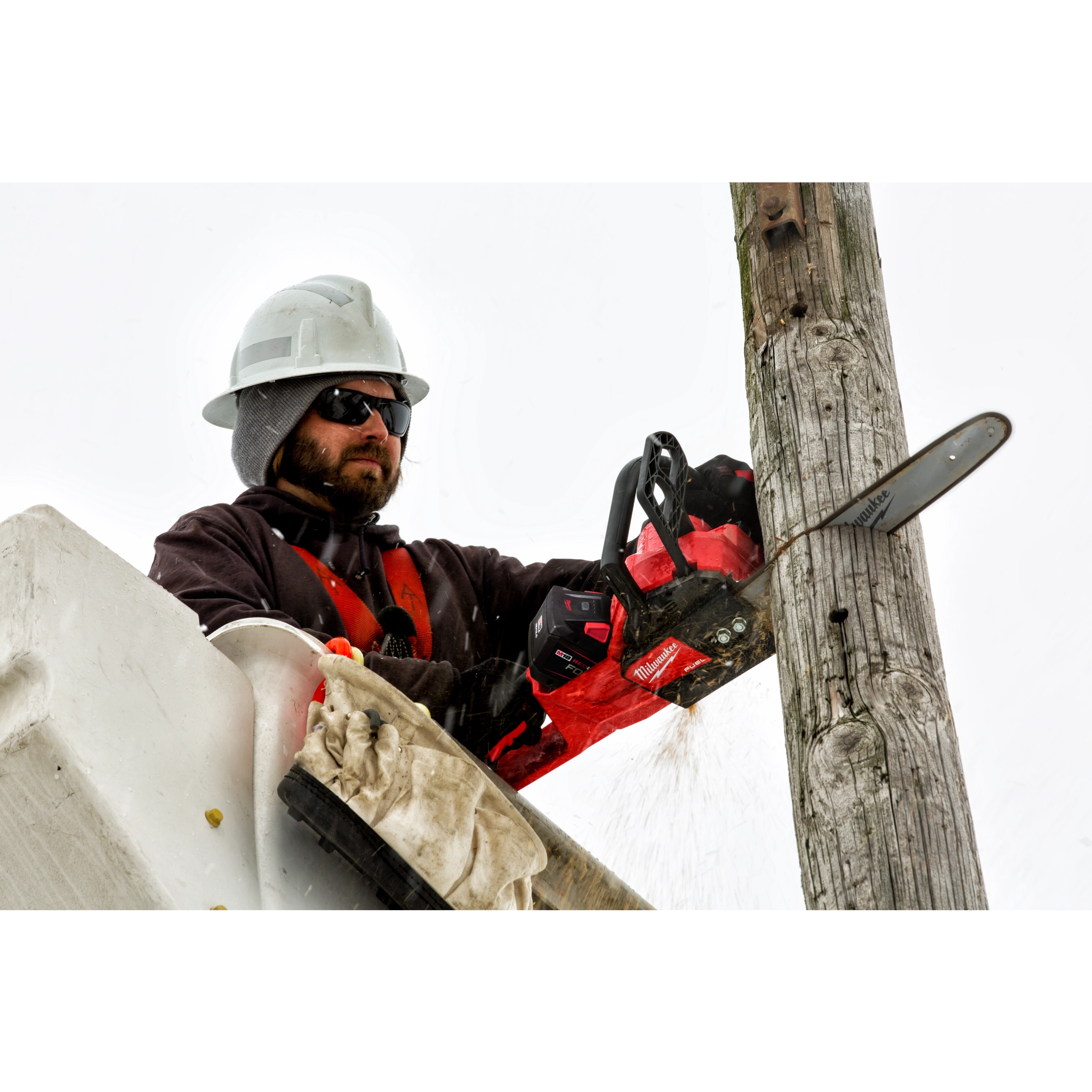 Person using a chainsaw to cut a wooden utility pole while wearing protective gear and a hard hat.