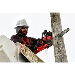 Person using a chainsaw to cut a wooden utility pole while wearing protective gear and a hard hat.