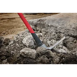 A 5lb Pick Mattock with a 36-inch handle is shown in use, breaking through hard soil and rocks. The tool's red and black handle is visible, along with the pick head firmly lodged into the ground, demonstrating its capability for heavy-duty digging and excavation tasks.