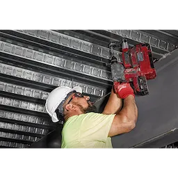 Worker wearing a hard hat and safety gloves uses a red power drill on a metal structure. Ear plugs are recommended for noise protection.