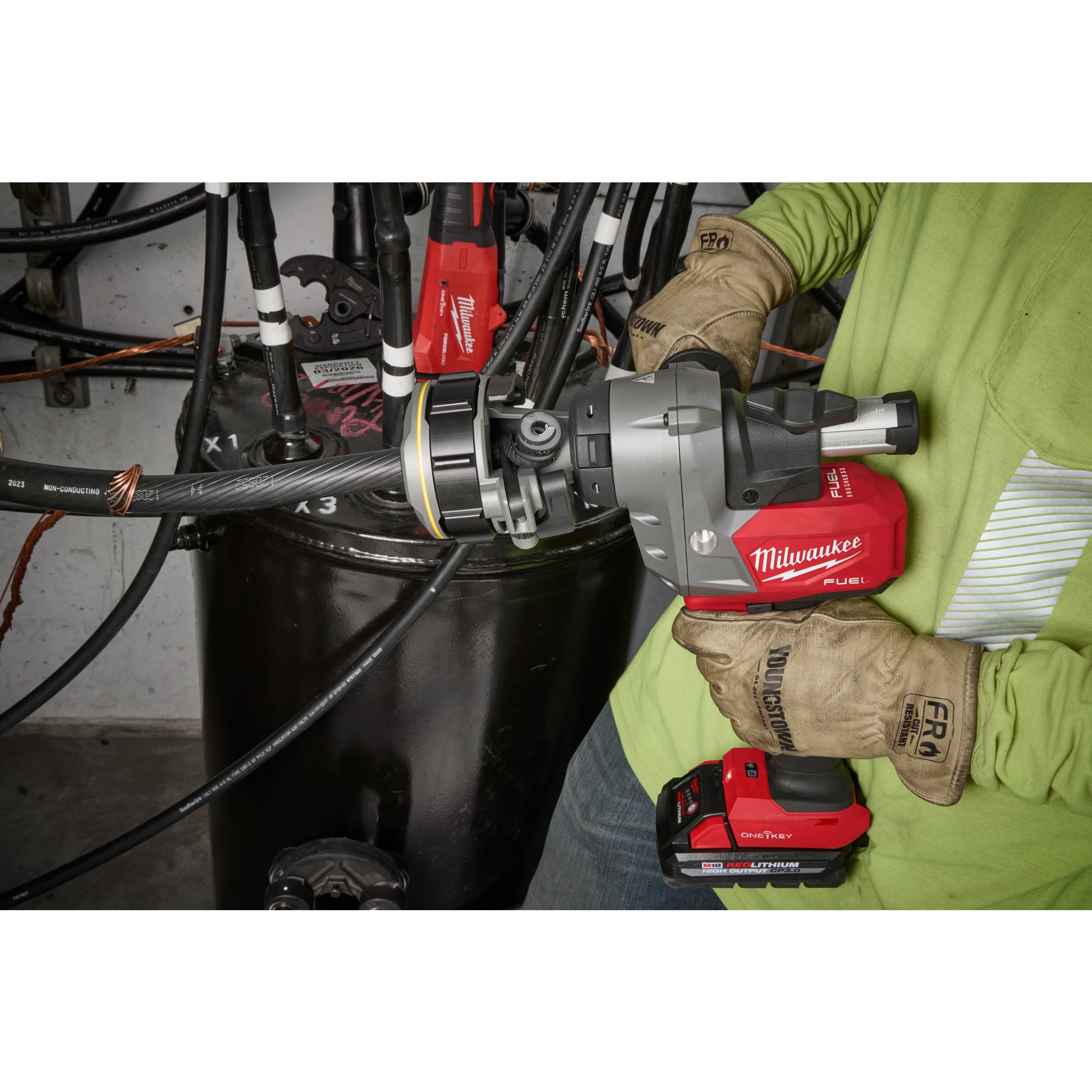 A person wearing gloves operates an M18 FUEL™ Covered Conductor Cable Stripper from Milwaukee Tool, stripping a cable in an industrial setting. The red and black power tool is seen in use, efficiently removing the cable's outer layer.