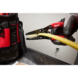 Gloved hand using the 8-20 AWG Dipped Grip Wire Stripper & Cutter (USA) to strip the insulation from a yellow wire, with a tool bag nearby.