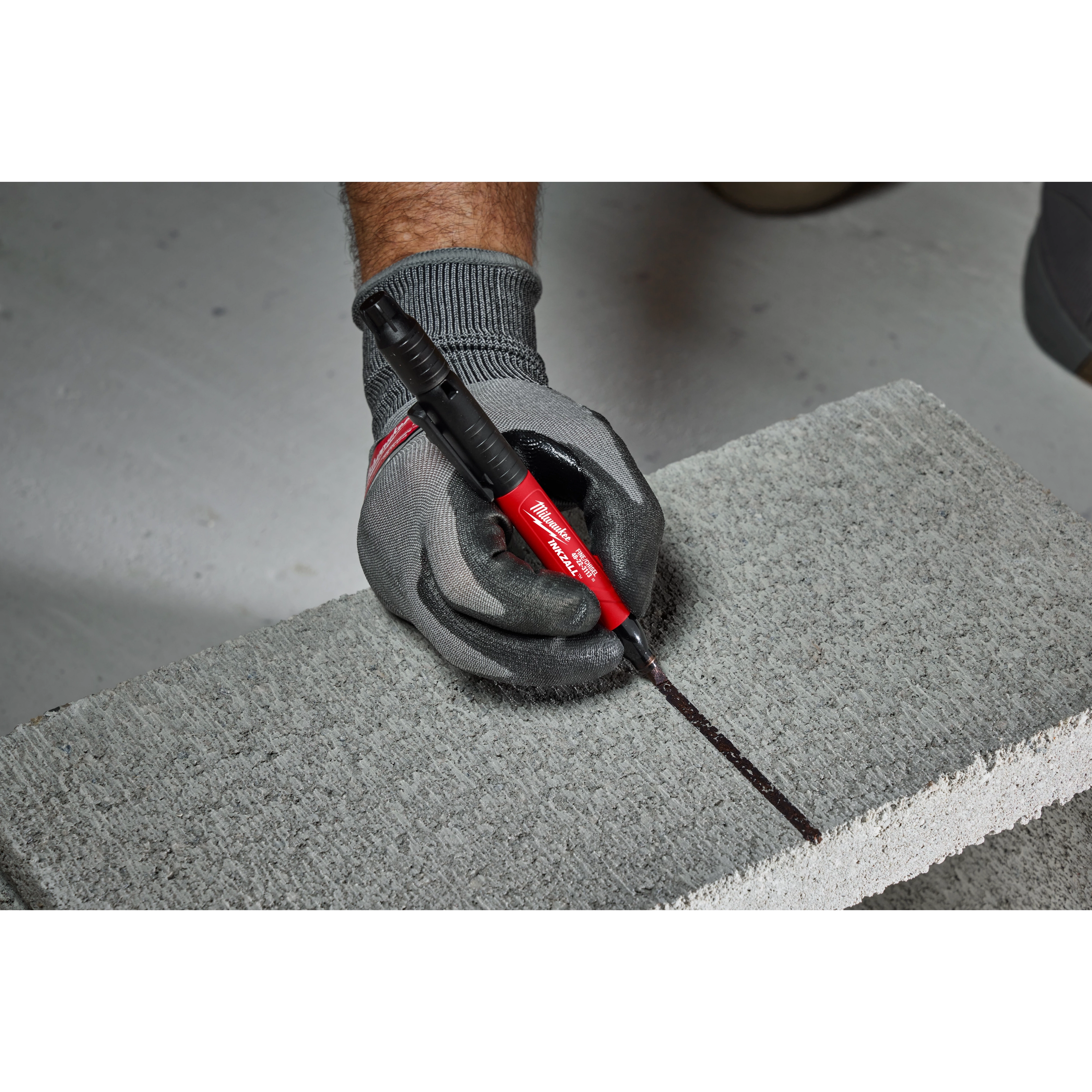 A person wearing gray gloves uses the INKZALL™ Double Sided Black Jobsite Marker – Ultra Fine & Fine Point to draw a black line on a concrete block. The marker has a red body and black dual tips for precision marking on various surfaces.