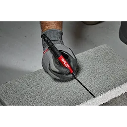 A person wearing gray gloves uses the INKZALL™ Double Sided Black Jobsite Marker – Ultra Fine & Fine Point to draw a black line on a concrete block. The marker has a red body and black dual tips for precision marking on various surfaces.
