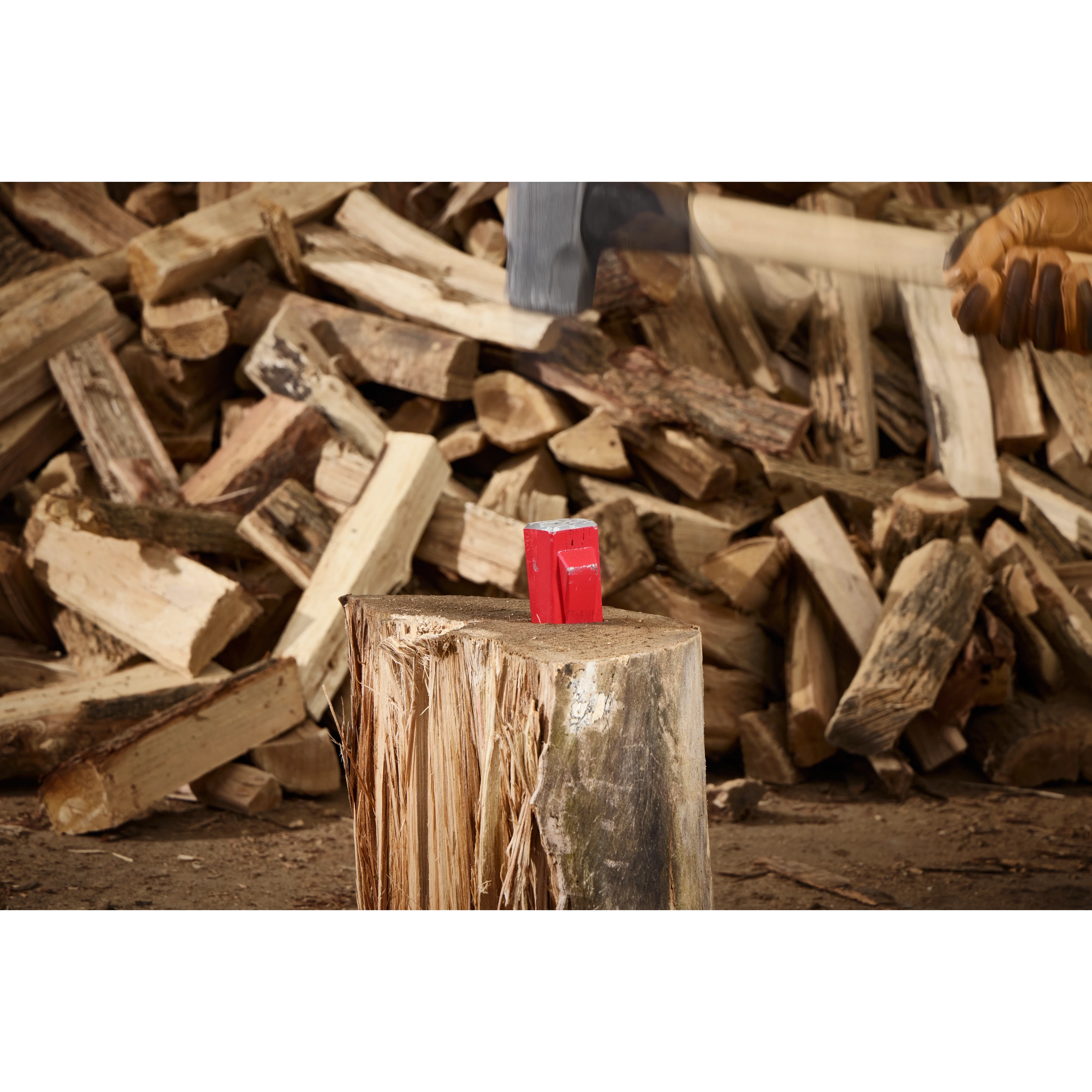 The 5lb Splitting Wedge is placed on a large log ready for splitting, surrounded by stacks of chopped firewood. A hand wearing a work glove holds an axe poised above the wedge, about to strike. The scene is set in a woodworking area.