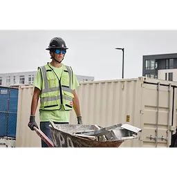A construction worker wearing a green safety vest, helmet, and gloves holds a wheelbarrow filled with metal debris. The worker is wearing Full Frame Safety Glasses with Removable Side Shields – Blue Mirrored Anti-Scratch Lenses. The background includes shipping containers and buildings.