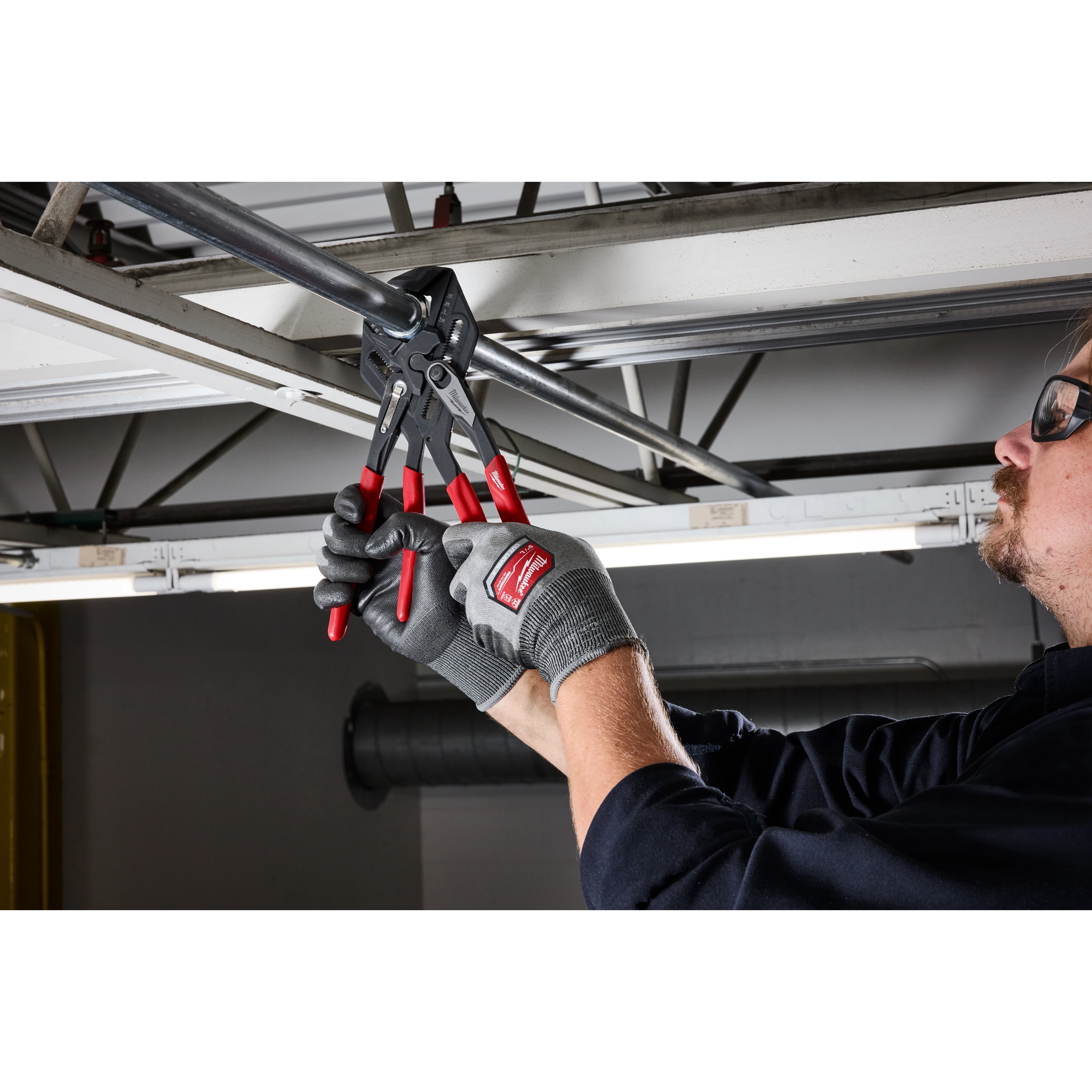 A person uses 10" Pliers Wrench with red handles to adjust a metal pipe positioned on a ceiling beam in a workshop area.