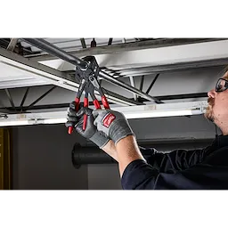 A person uses 10" Pliers Wrench with red handles to adjust a metal pipe positioned on a ceiling beam in a workshop area.