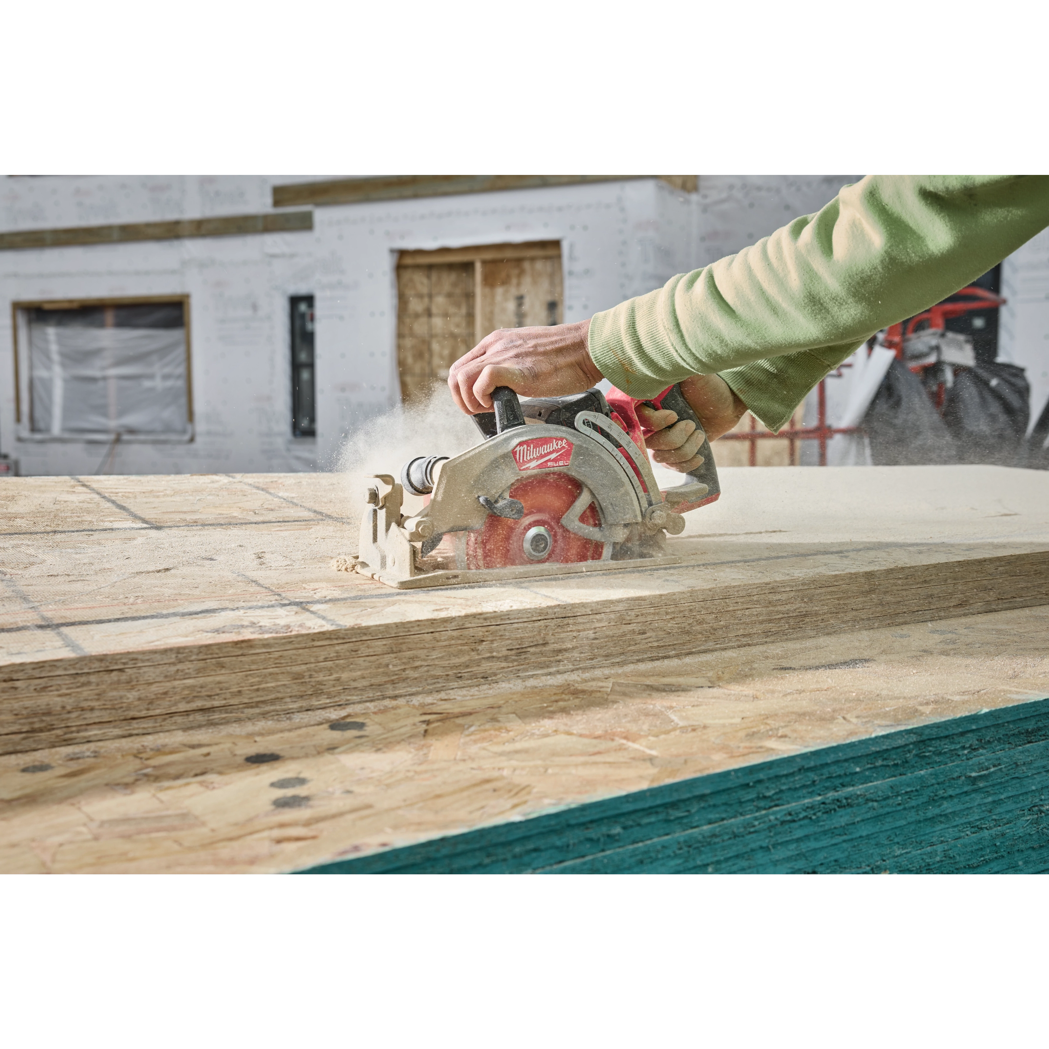 A person operates a M18 FUEL™ 7-1/4" Rear Handle Circular Saw to cut through wooden boards at a construction site. Dust is visible around the cutting area, indicating active use. A partially constructed building is in the background.