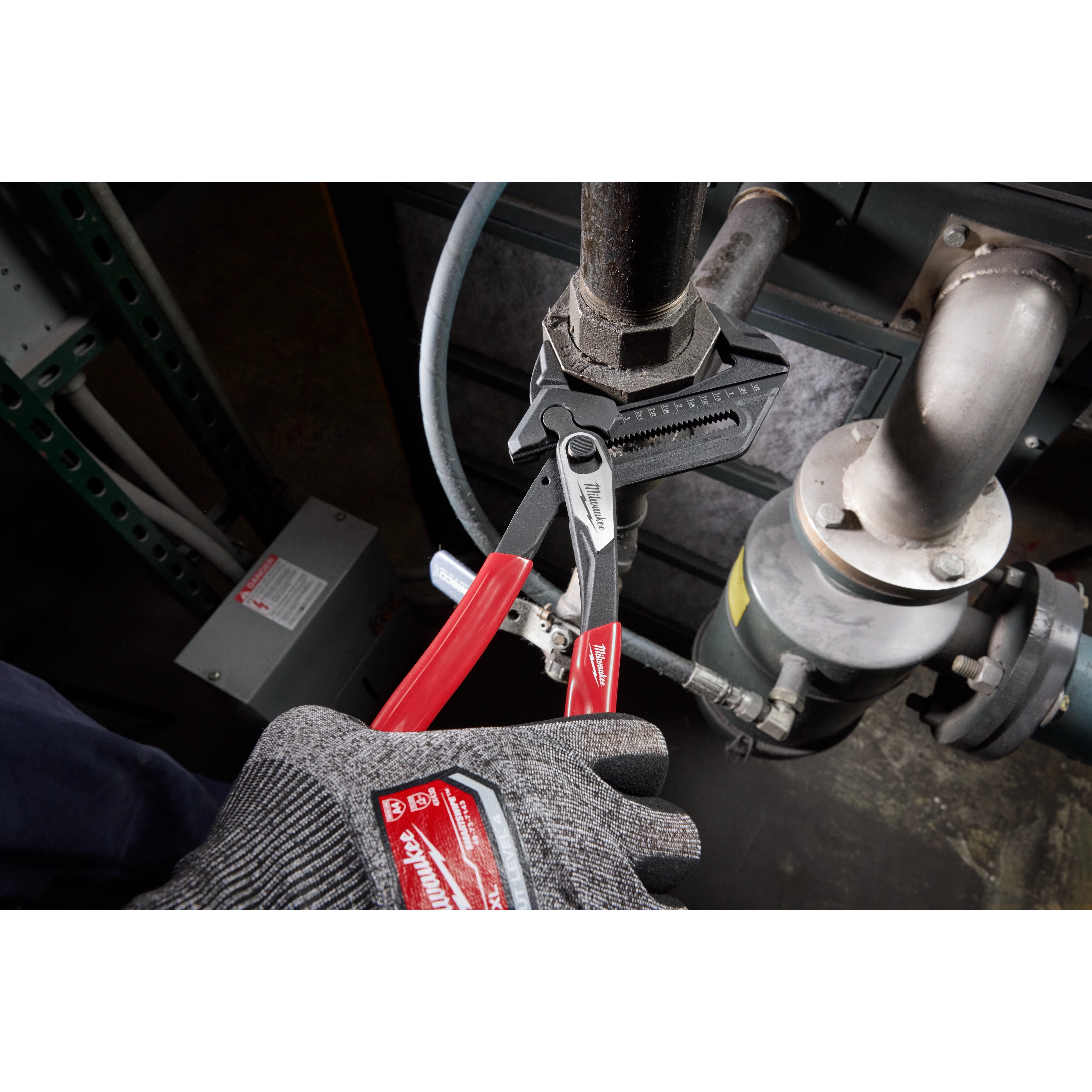 A gloved hand is using 12" Pliers Wrench with red handles to tighten a pipe fitting. The wrench has textured jaws for gripping.