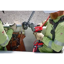 Two workers use the M18 FUEL™ Covered Conductor Cable Stripper to strip a cable. One worker holds the cable while the other operates the red and black handheld device. Both are wearing safety harnesses and green work shirts.