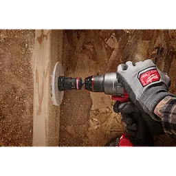 A person wearing gloves uses a power drill equipped with a 3/8" QUIK-LOK™ Arbor Set to drill a hole in a wooden surface. Wood dust is visible around the drill bit, and the background consists of unfinished wooden boards.