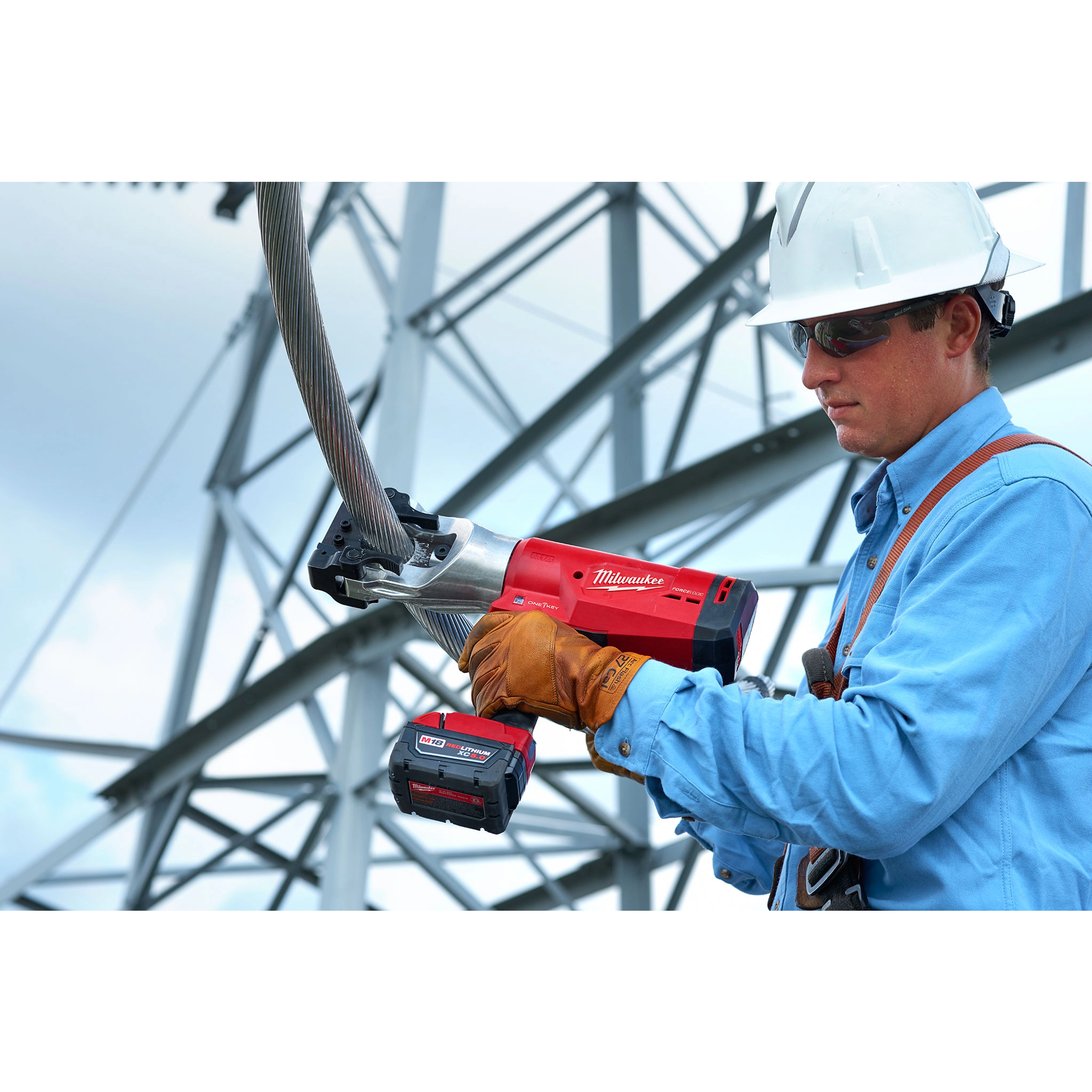 A worker wearing protective gear operates the FORCELOGIC™ M18™ 1590 ACSR Cutter Kit, cutting through a thick cable. The tool is red and black, battery-powered, and designed for efficient cable cutting, shown in an industrial setting with metal structures in the background.