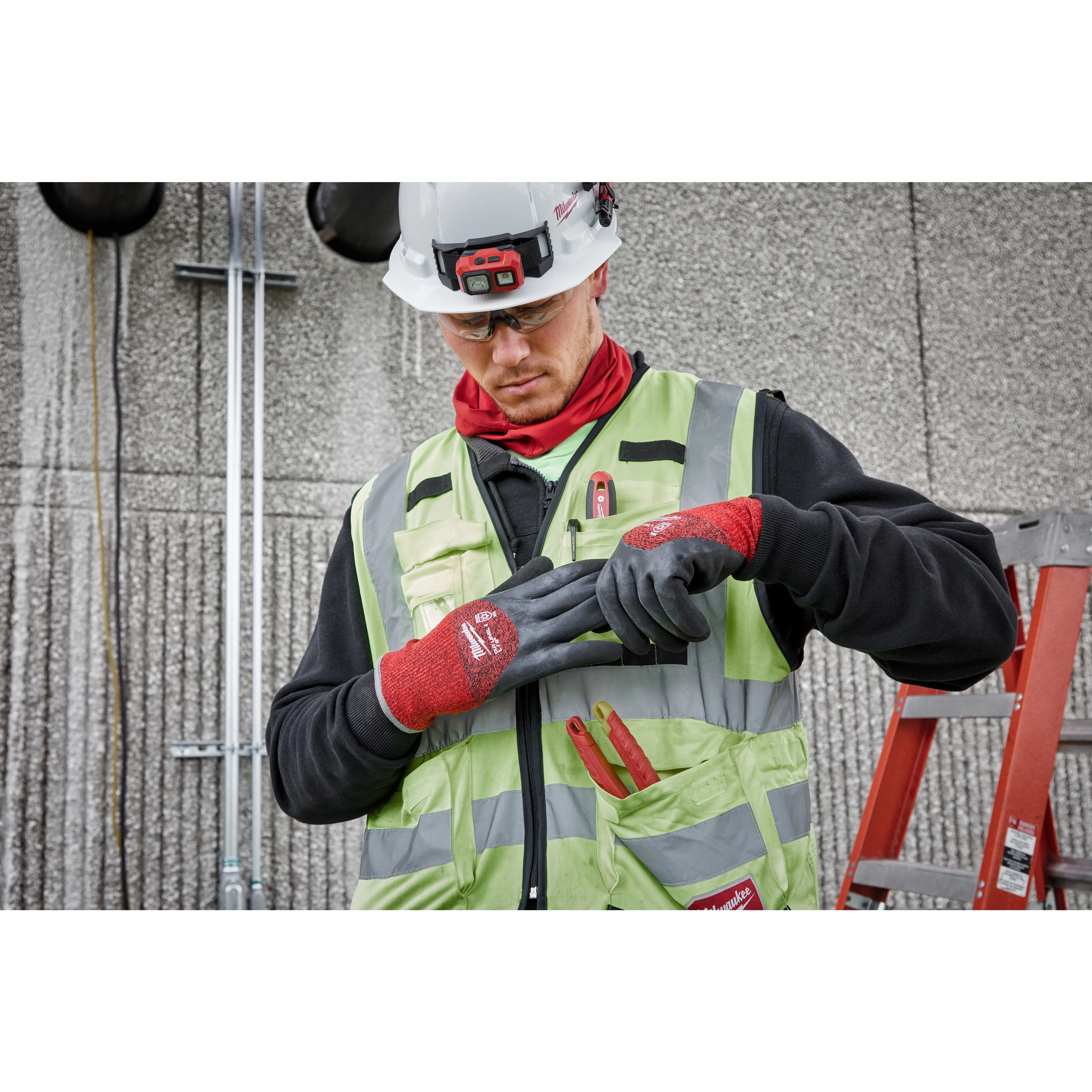 A construction worker wears a high-visibility vest and a hard hat while adjusting Cut Level 4 Winter Dipped Gloves - L. The gloves are red and black, designed for hand protection in cold conditions. The worker stands near a ladder and a wall with mounted equipment.
