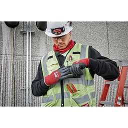 A construction worker wears a high-visibility vest and a hard hat while adjusting Cut Level 4 Winter Dipped Gloves - L. The gloves are red and black, designed for hand protection in cold conditions. The worker stands near a ladder and a wall with mounted equipment.