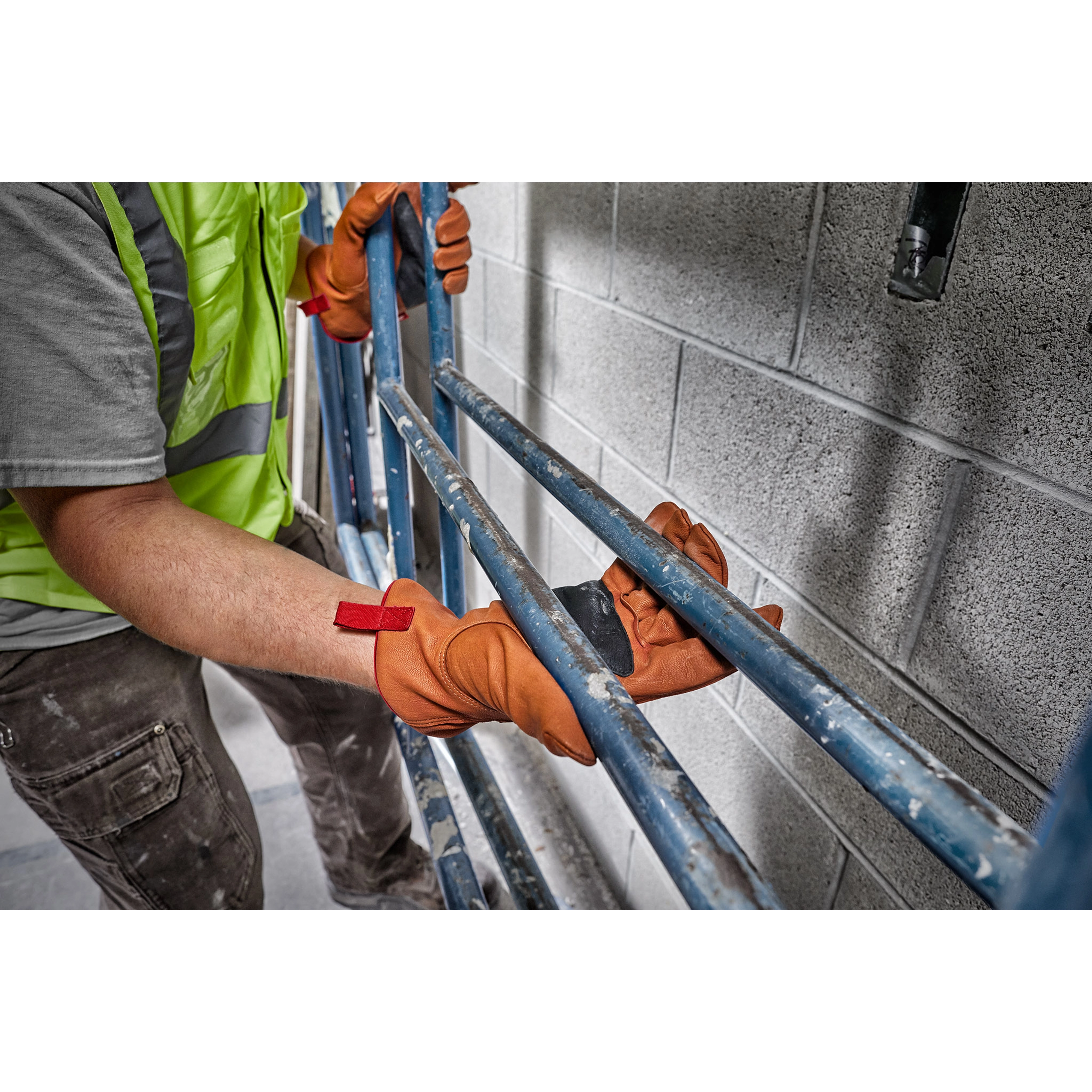 Worker wearing Goatskin Leather Gloves handling blue metal scaffolding. Worker wears a yellow safety vest and grey shirt.