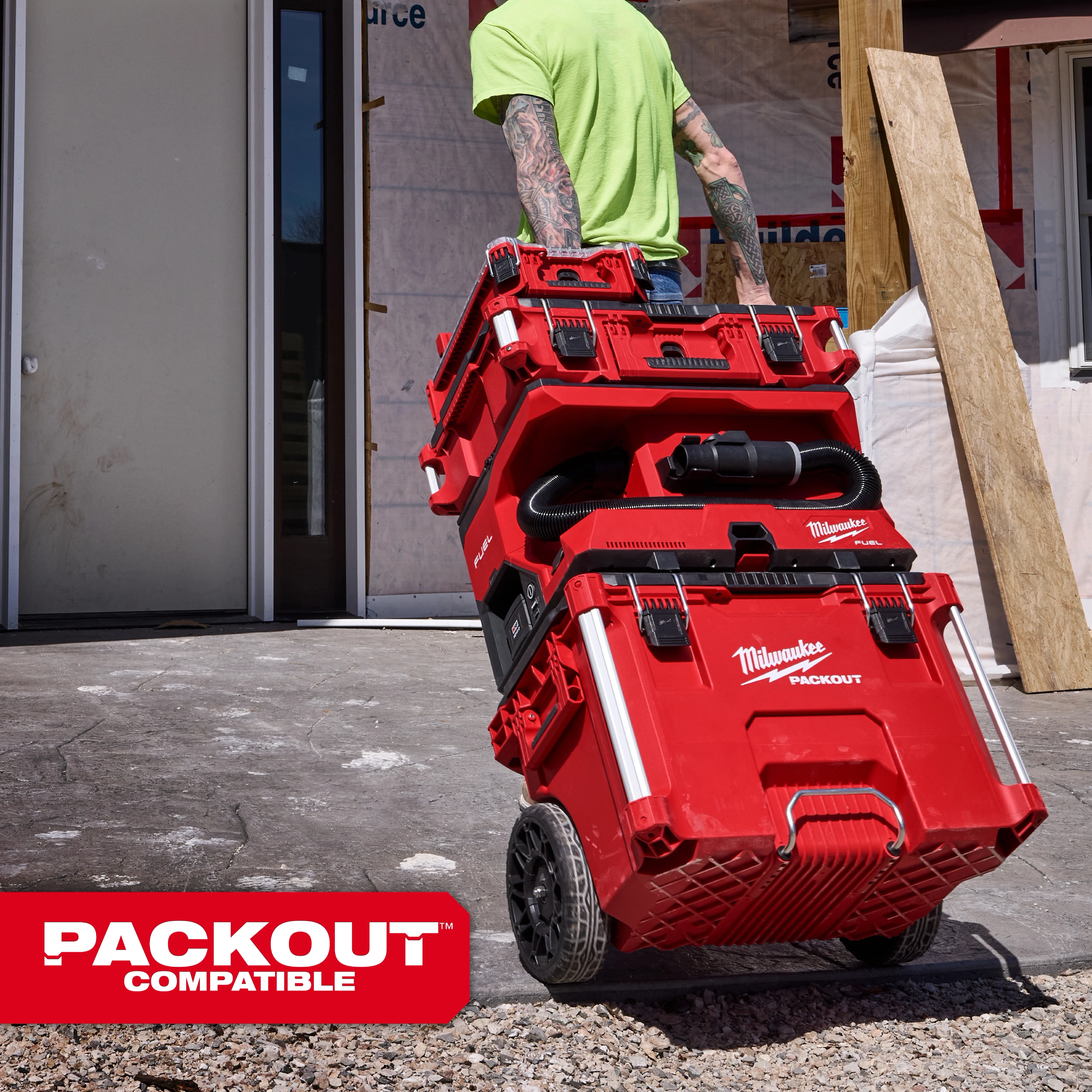 Worker pulls stacked red M18 FUEL 4 Gallon Wet/Dry Vacuum w/ PACKOUT Compatibility & VACLINK on wheels at a jobsite.