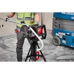 A worker operates the Milwaukee M18 FUEL Pipe Bender, which is mounted on a tripod stand. The device is bending metal piping in a construction setting. The worker wears a green safety vest and grey gloves, with additional pipes and construction equipment visible in the background.