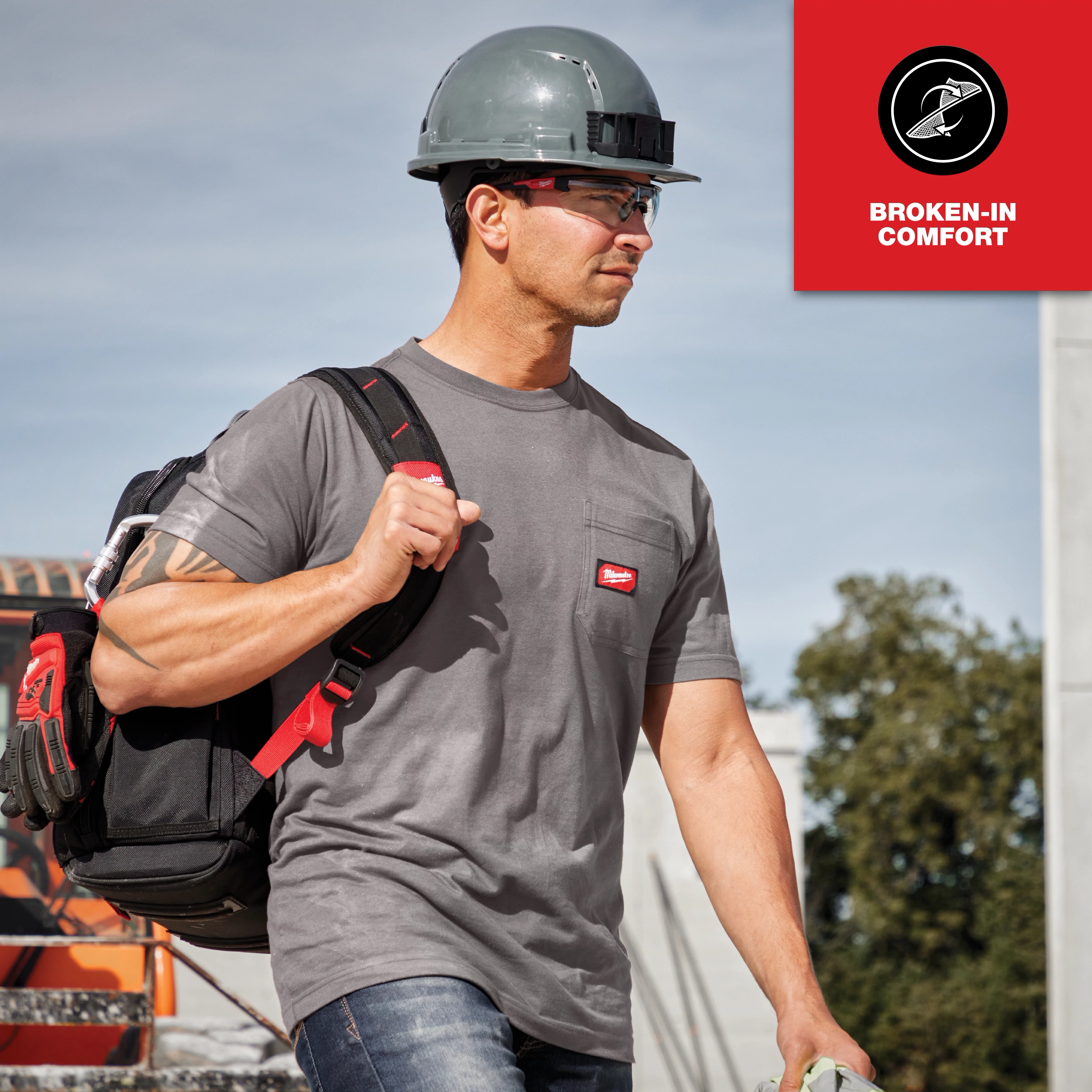 Man wearing a gray GRIDIRON Pocket T-Shirt - Short Sleeve and a hard hat carries a backpack at a construction site. "Broken-in comfort" text is seen.