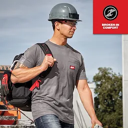 Man wearing a gray GRIDIRON Pocket T-Shirt - Short Sleeve and a hard hat carries a backpack at a construction site. "Broken-in comfort" text is seen.