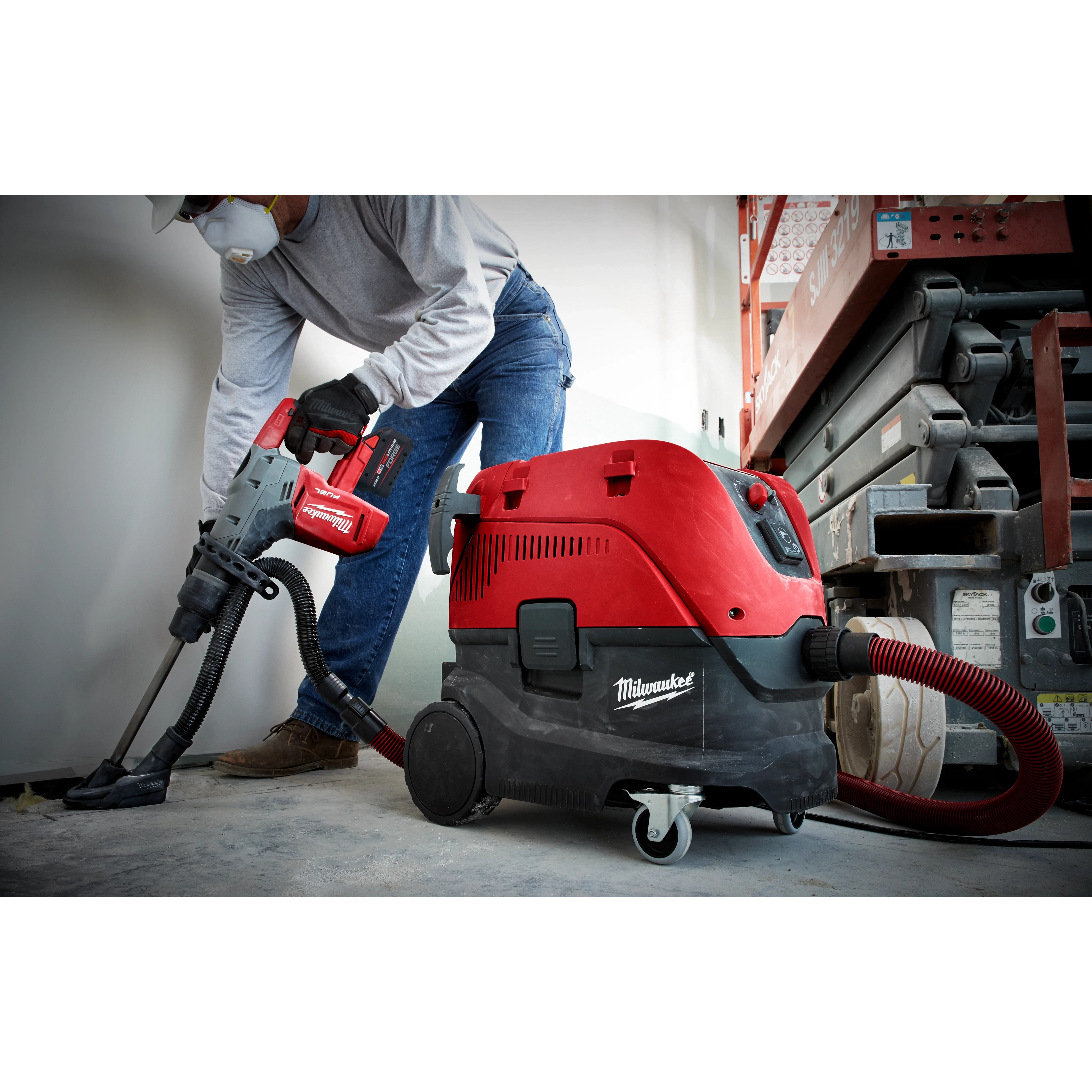 A person operates the M18 FUEL™ Rotary Hammer with Chisel Boot, M18 FUEL 1-9/16 SDS MAX Hammer Drill. The tool is red and black and is connected to a Milwaukee vacuum system for dust collection. The person wears safety gear and stands next to a construction lift.