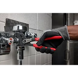 A person wearing gloves uses the 10" Pliers Wrench to tighten a metal pipe fixture in a tiled bathroom. The wrench has black jaws and red handles.