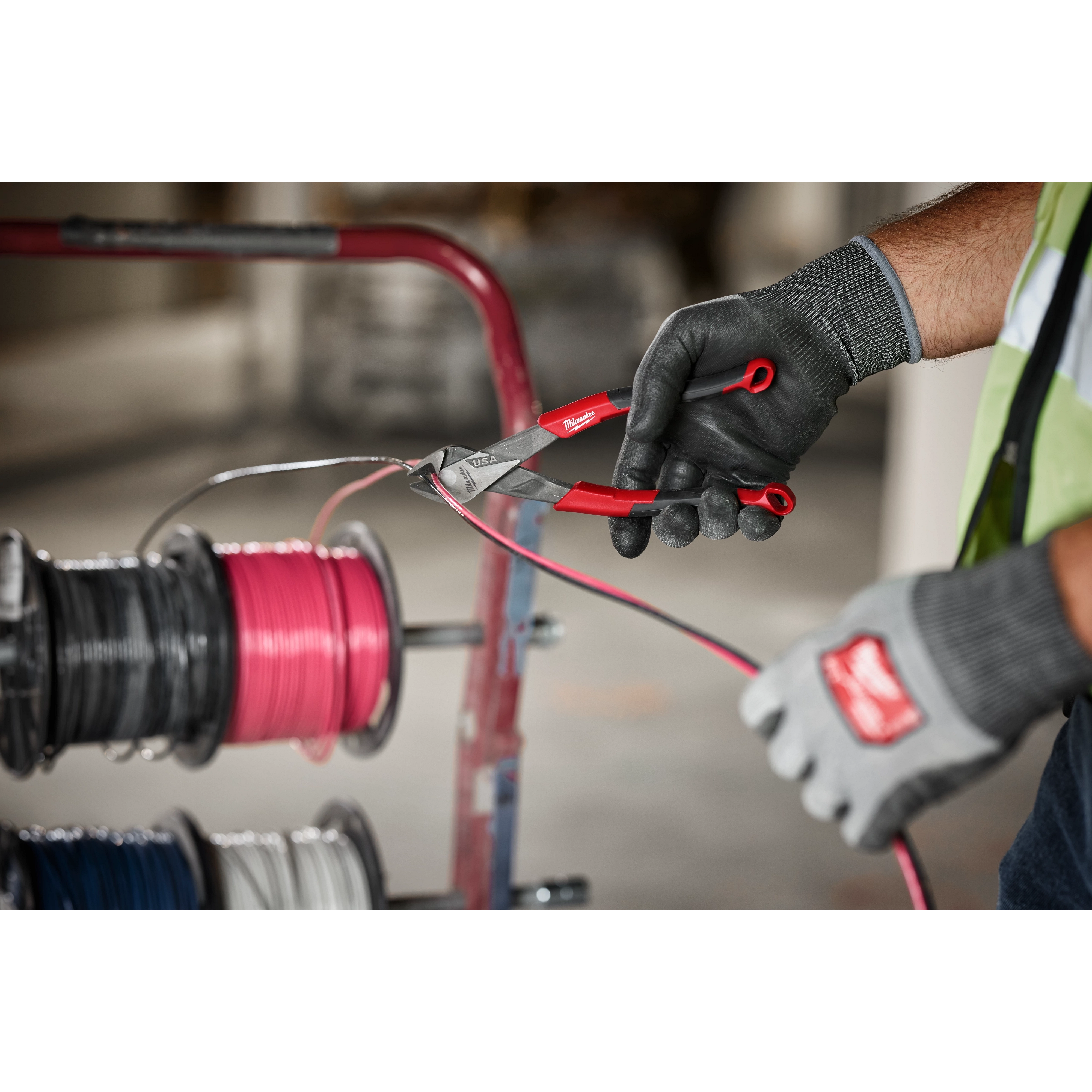 A person in work gloves uses the 8" Diagonal Comfort Grip Pliers (USA) to cut a pink wire, with wire spools in the background.