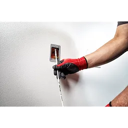 A person wearing red and black gloves uses the 5' Mid Flex Fish Stick to pull wires through an electrical outlet box in a wall.