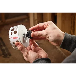 A person is assembling a tool from the 10PC QUIK-LOK™ BIG HAWG™ PACKOUT™ Kit, holding a white metal hole saw labeled "HOLE DOZER" with another part being attached to it, in an indoor workshop setting.