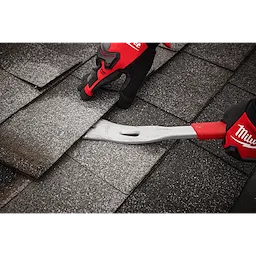 The 16" Molding Puller from Milwaukee is being used to remove roof shingles. A person wearing protective gloves grips the metal tool with a red handle as they lift the edge of the shingles, revealing the layer beneath. There are multiple shingles visible in the background.