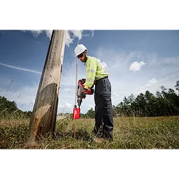 A construction worker uses the Milwaukee SDS Max Side Load Ground Rod Driver to drive a grounding rod into the earth next to a wooden utility pole in an open field.