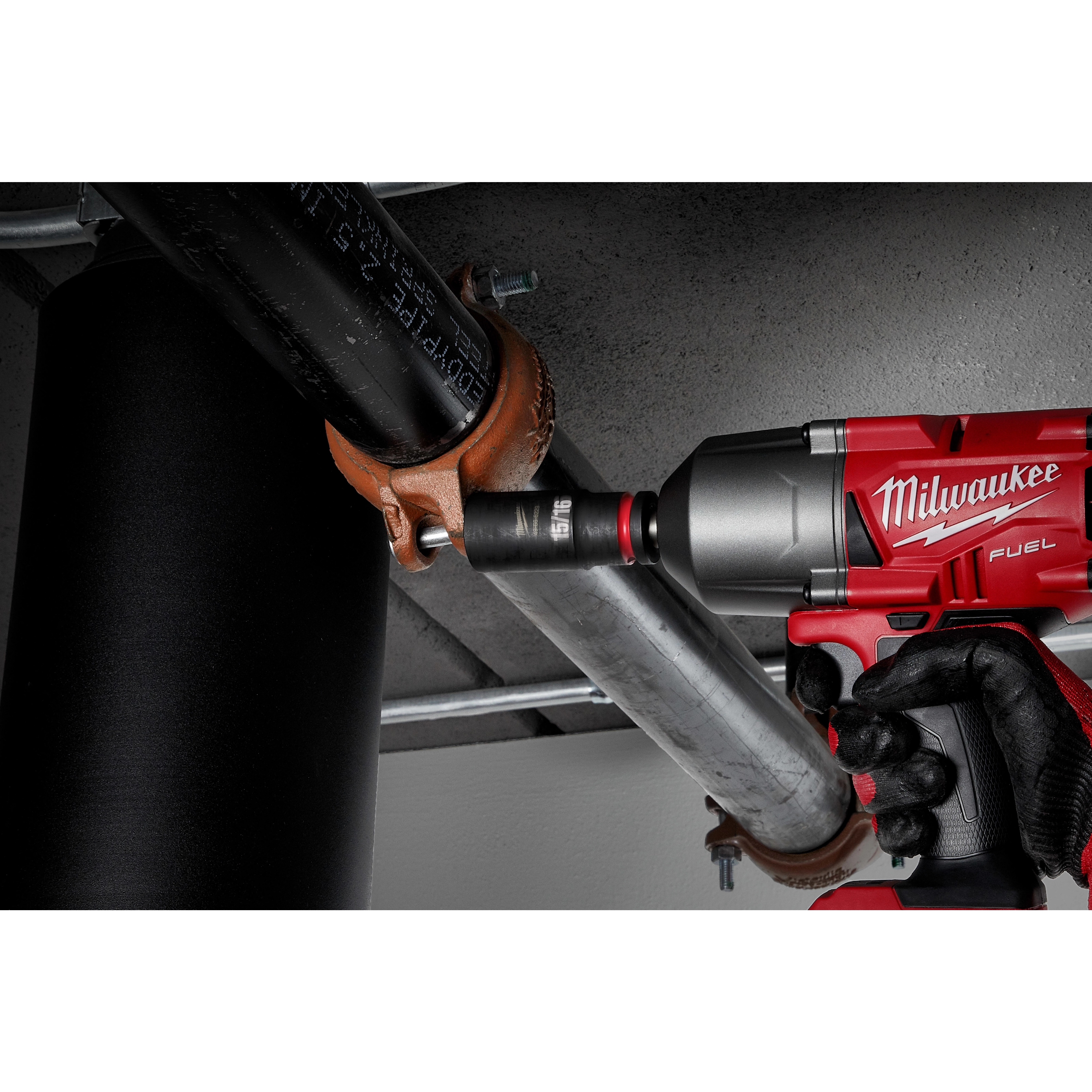 A worker uses a red Milwaukee power tool equipped with a SHOCKWAVE Impact Duty™ 1/2" Drive 15/16" Deep 6 Point Socket to fasten a pipe clamp onto a metal pipe. The setting appears to be an industrial or construction environment.