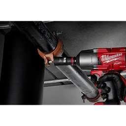 A worker uses a red Milwaukee power tool equipped with a SHOCKWAVE Impact Duty™ 1/2" Drive 15/16" Deep 6 Point Socket to fasten a pipe clamp onto a metal pipe. The setting appears to be an industrial or construction environment.